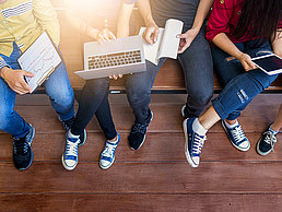 Vier studierende sitzen auf einer Bank mit Laptop, Tablet und Block in der Hand.